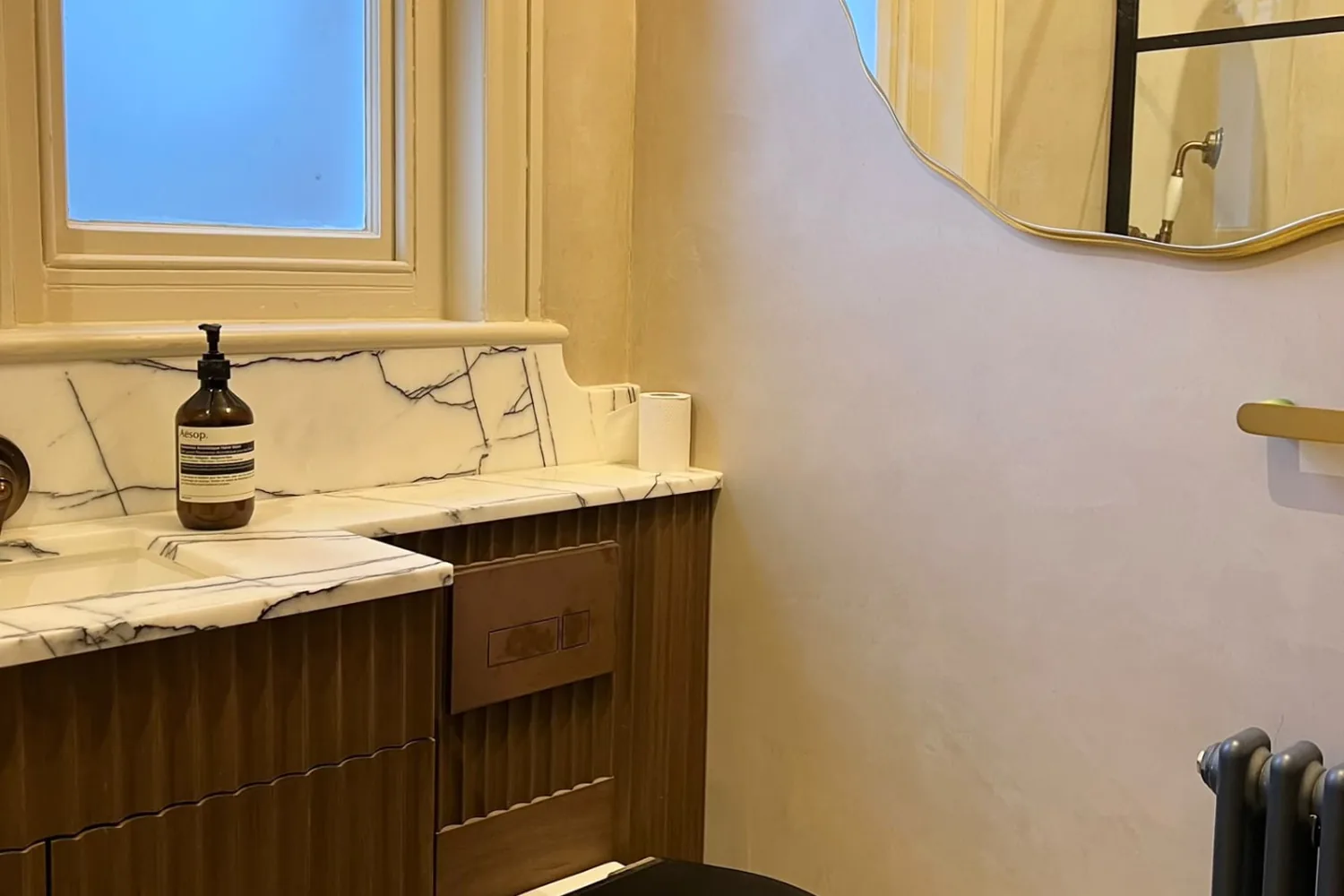 Warm microcement bathroom walls beside a marble worktop, wavy mirror and shower screen